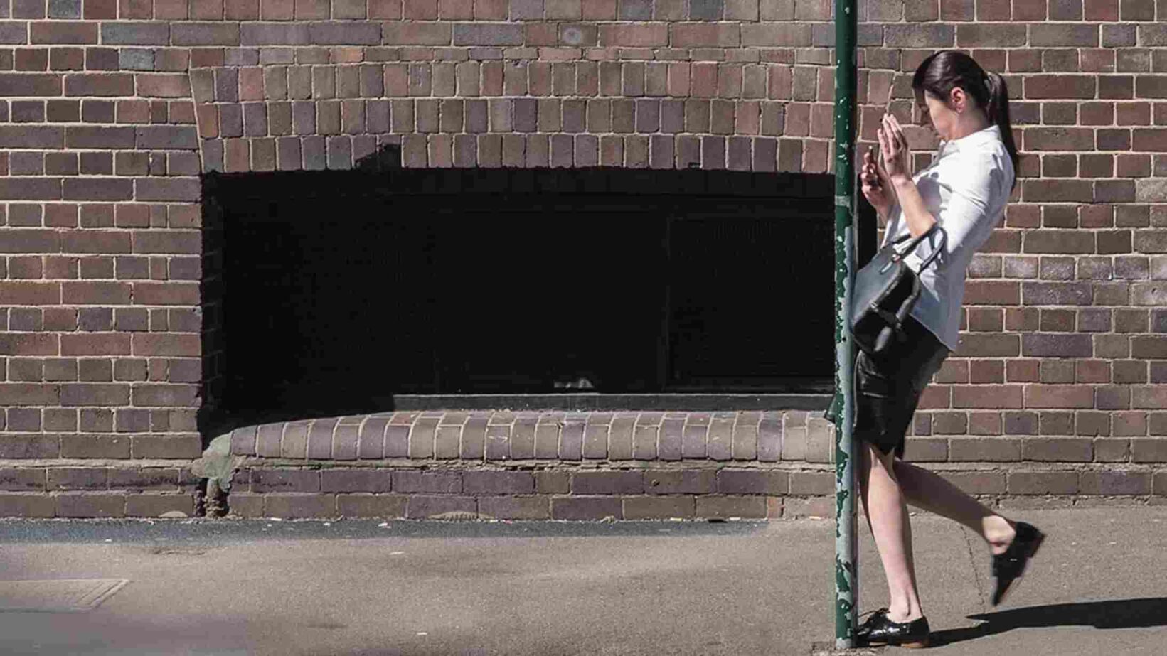 Urban professional moment captured by reborn digital marketing agency - reborn - digital creative agency in sydney A professional woman stands next to a brick wall, leaning against a pole and using her phone, creating a casual yet sophisticated moment. The image conveys a sense of modern urban life, possibly featured in a lifestyle or business campaign managed by Reborn Digital Marketing Agency.