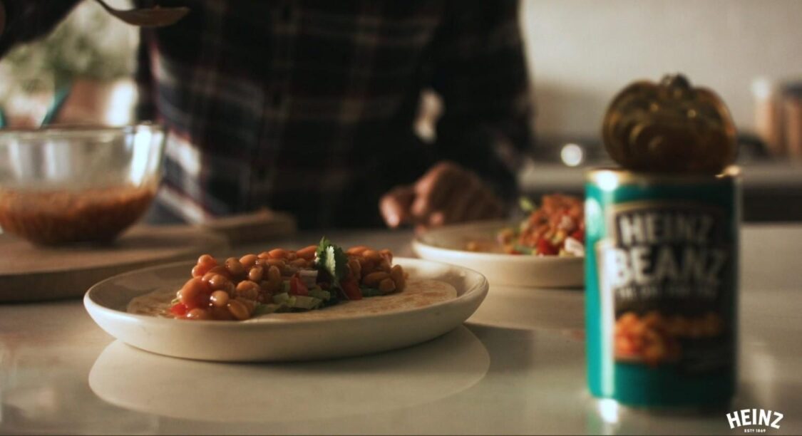 A person prepares a meal, spooning Heinz baked beans onto a Heinz Beanz being served on a tortilla with fresh toppings, part of a creative food advertising campaign