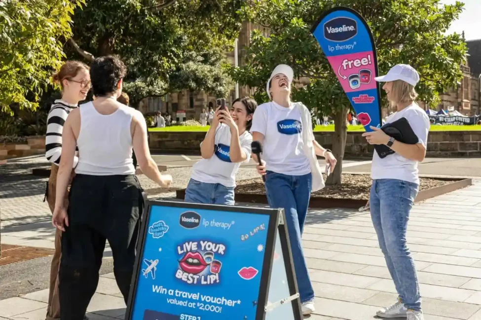 A group of people outdoors, two wearing Vaseline shirts, smile and interact animatedly near a sign promoting lip therapy and a travel voucher giveaway.